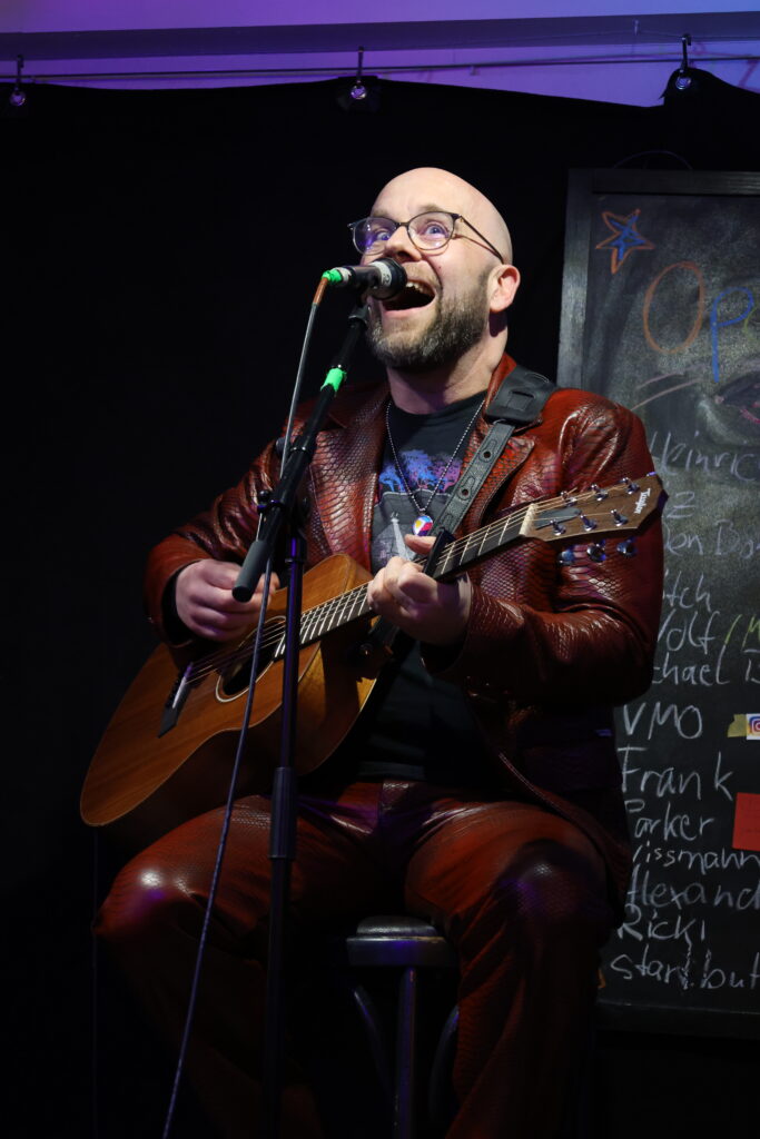 Michael Batisch singt fröhlich und spielt Gitarre auf einer Bühne